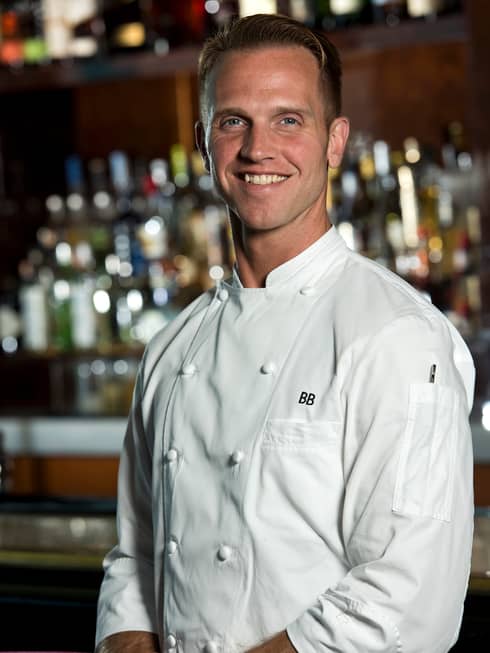 Portrait photo of Executive Chef Brian Becher in white uniform, smiling and standing in front of bar