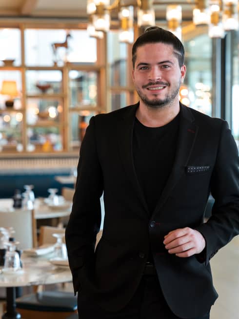 A man in a fitted black jacket stands in a well-lit dining room