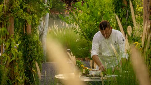 A chef works at an outdoor wooden table surrounded by gardens