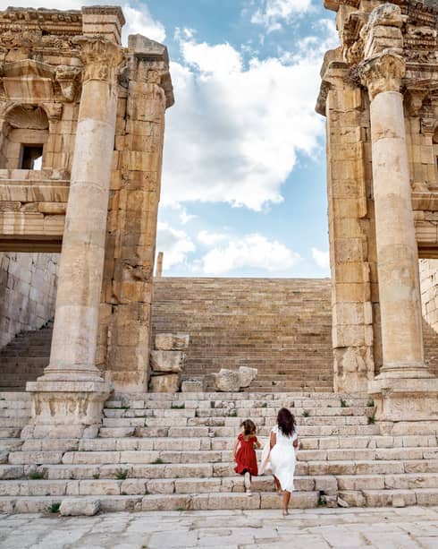 Rear view of an adult and child climbing the column-lined steps of a stone ruin toward a bright blue, cloud-steaked sky.