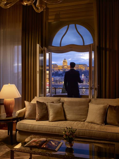 A person standing on a balcony at dusk, holding a glass of champagne, overlooking a lit historic building.