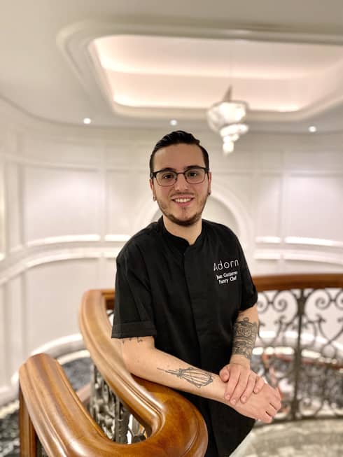 A young male pastry chef with a few tattoos leaning on a railing in a large stairwell.