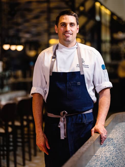 A male chef in an apron standing next to a counter in a dimly lit restaurant.