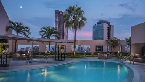 Rooftop pool at dusk with wicker furniture, palm trees, lights, skyline in background