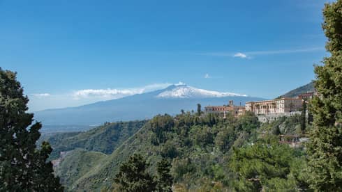 View from afar of an expansive mediterranean villa atop a forested hill, snow-capped mountain in the distance.   