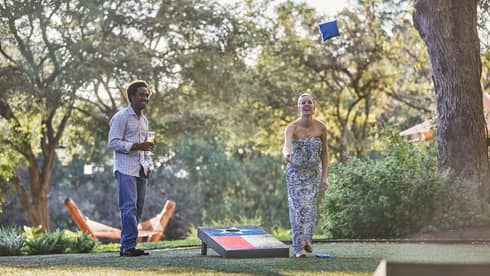 Laughing woman tosses bean bag on lawn, man holding beer watches