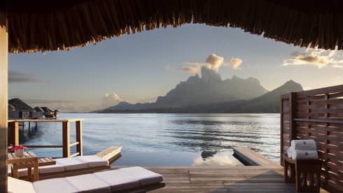 A view from a wooden Water Bungalow with white cushioned lounge chairs overlooking the ocean and mountains in the background at Four Seasons Bora Bora