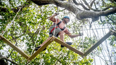 A man wearing a red shirt and grey shorts is climbing through an aerial ropes course