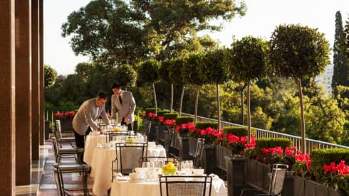 Two servers in grey jackets with black ties set outdoor dining tables with white tablecloths, dishes, red flowers along side