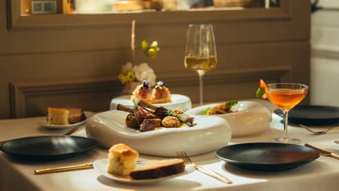 Three white serving platters with food sit in the middle of a white-cloth dining table with three black plates, a glass of white wine, a cocktail in a coupe and two small plates of bread surrounding them