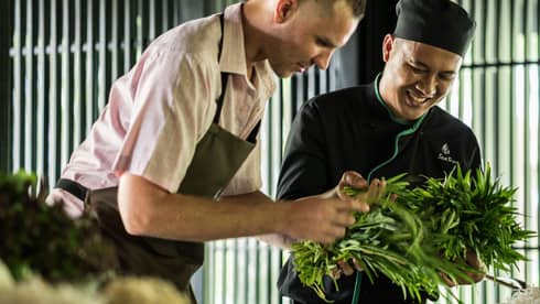 Smiling Chef Tran Van Sen holds greens, shows them to man in apron