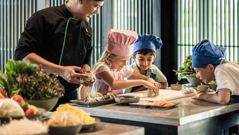 Chef watches as children wearing tall chef hats make spring rolls at counter