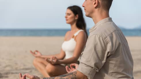 Two people meditating in a cross-legged position on a sandy beach, with the ocean in the background.