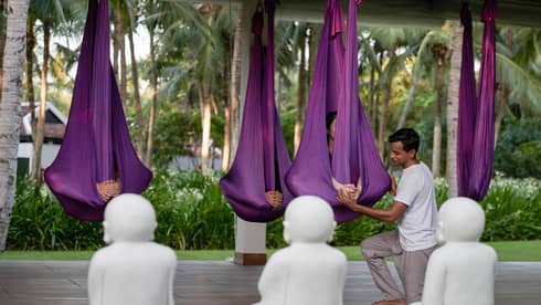 An instructor assisting participants during an outdoor aerial yoga session, with people suspended in purple hammocks surrounded by tropical greenery and white Buddha statues.