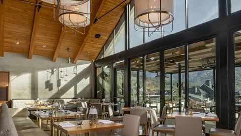 Modern restaurant interior with high wooden ceilings, large windows, and natural light. Tables are set, and an outdoor terrace offers views of surrounding hills.
