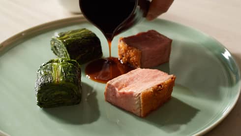 Close-up of squares of pork with crispy skin and rolls of cooked cabbage. A sauce is gently poured in the center of the food.