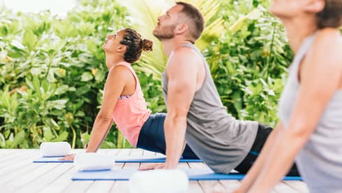 Three people each laying on a blue yoga mat with arms extended and faces looking upward are lined up in a row on an outdoor deck with greenery in the background