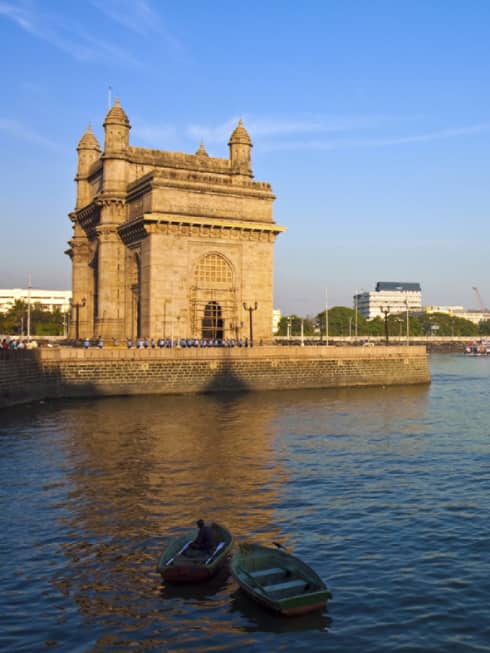 Harbourside view of an imposing towered stone structure, scattered rowboats in the foreground, larger moored ships beyond.