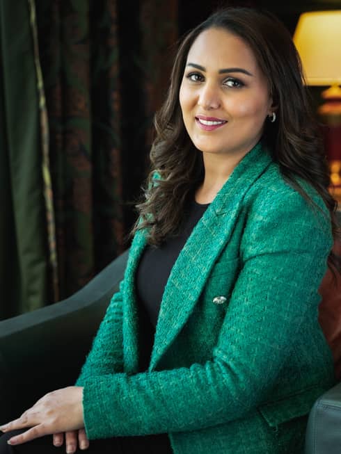 Woman with long dark hair wearing bright green tweed blazer, black shirt and black pants sits on an armchair and smiles at the camera