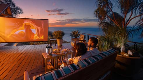 Two people sit on an outdoor sofa with a small table of food infront of them as they watch a movie on a large outdoor screen set up on a private terrace
