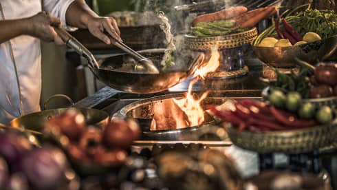 Close-up of chef stirring meals in copper pans over stove, flames 