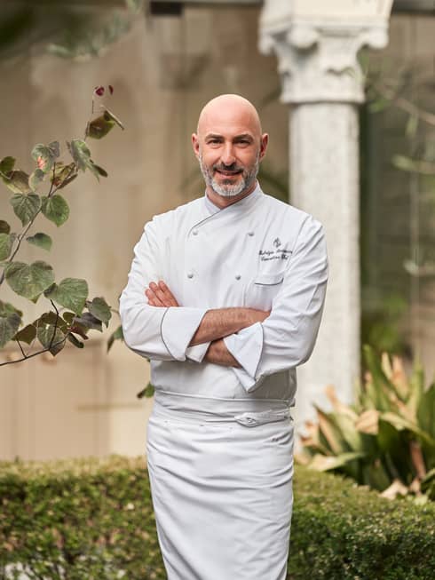 A man in chefs clothes with a bald head and a short beard is standing outside near plants and stone walls.