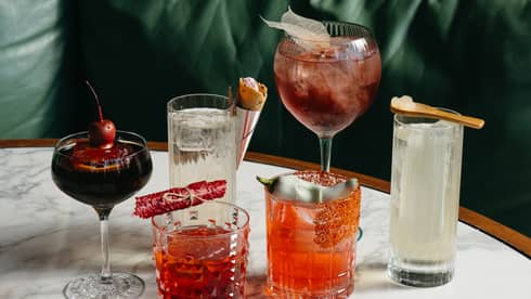 Six glasses and wine glasses with various coloured drinks placed beside one another on a dining table.