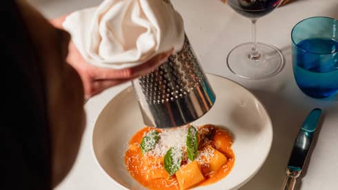 Person grating cheese over a pasta dish with a wine glass and silverware on the side