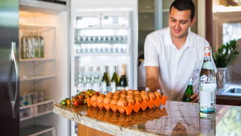 Smiling man holds green bottle in front of open fridge, fresh eggs on counter