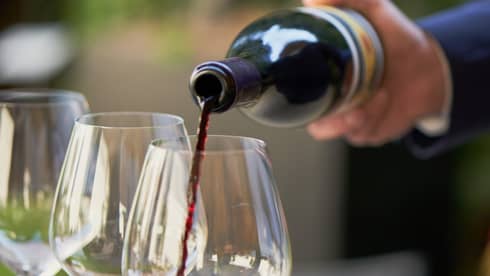 A close up of hands pouring a bottle of red wine into a clear wine glass that rests on a wooden tray, outdoors