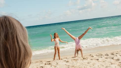 Two girls do cartwheels on beach as woman sits under an umbrella