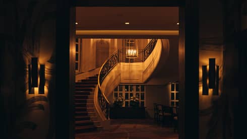 An interior space in the warm, low light of evening, featuring wall sconce lighting and a curved staircase and chandelier seen through a doorway.
