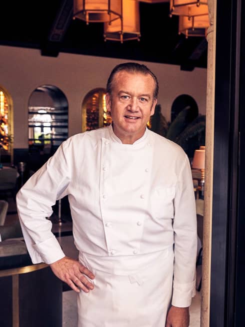 A male chef in white chef clothes standing in the doorway to a dimly lit restaurant.