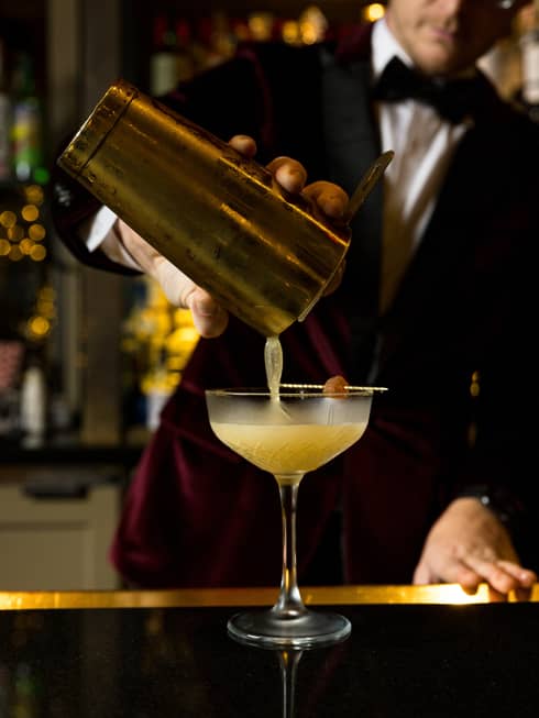 Bartender wearing a burgundy velvet suit and bow tie pours a cocktail from a shaker into a stemmed glass