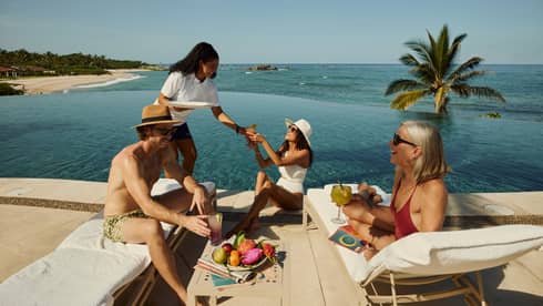 Three people mingle over cocktails next to an ocean-view resort pool, as server serves a drink
