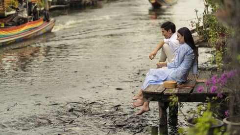 Couple sits on the dock of the Chao Phraya River, hungry fish swarming beneath their dangling feet