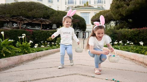 Two children in matching outfits wearing pink rabbit ears and holding Easter baskets with grand white building in backdrop