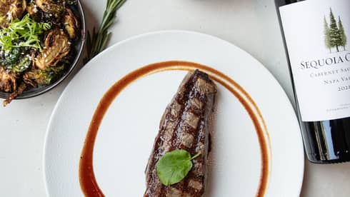 Strip steak on a white dish, bottle of wine, a small bowl of salt, small bowl of peppercorns, two bowls of food and a glass of wine on a white table