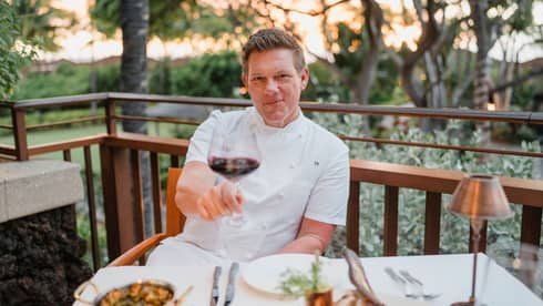 A smiling Tyler Florence sitting out on a veranda wearing his chef jacket and giving a toast with a glass of red wine.