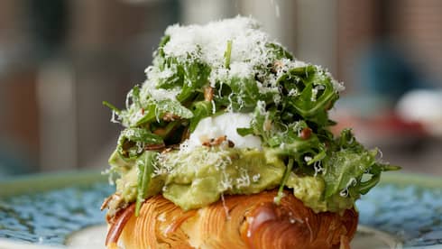 A close-up of a plate holding a croissant covered with avocado, egg and arugula salad topped with grated parmesan. 