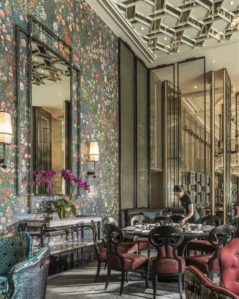 Hotel staff sets table with elegant red velvet chairs under tall mirror, floral wallpaper 