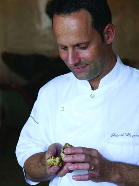 Portrait photo of Executive Chef Pascal Meynard wearing white chef's uniform, looking down at mushrooms in hands