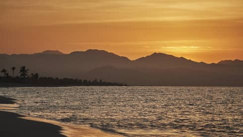 Beach and ocean in Los Cabos, Mexico, with sun setting behind distant mountains.