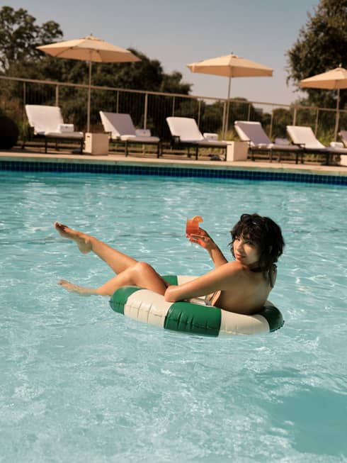 Guests lounges on a green-and-white striped pool float in the water while holding a cocktail in one hand