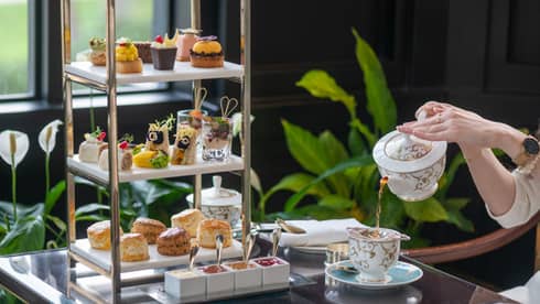 Three-tiered, chrome-edged display holds a variety of small desserts and baked goods set next to a person pouring tea from a pot into a gold-and-white tea cup