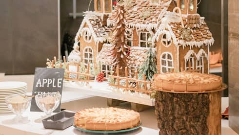 Extravant gingerbread house with three steeples and various trees and decor sits at the center of a table next to two sliced pies