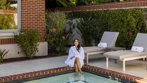 A woman dressed in a white spa robe sits on the corner of an outdoor pool surrounded by lush greenery