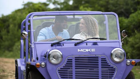 Front view of a couple driving a purple Moke, a small open-air recreational vehicle, along a dirt road surrounded by trees.