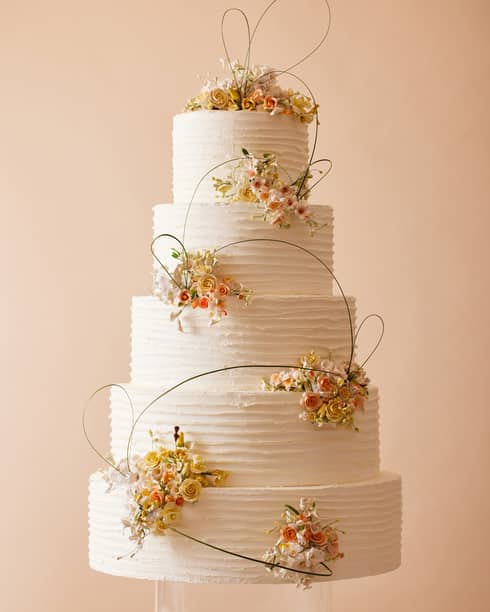 Five-tiered white wedding cake with bunches of fondant flowers in light orange and pinks alternating on each layer