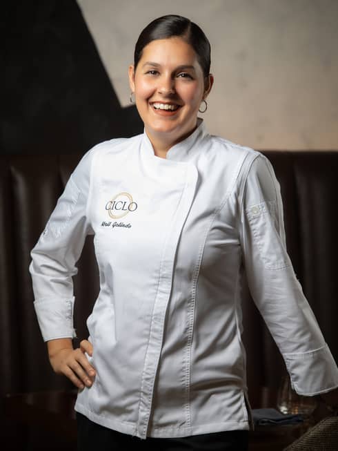 A portrait of female Chef Abril Galindo leaning against a table and smiling in her white chef coat